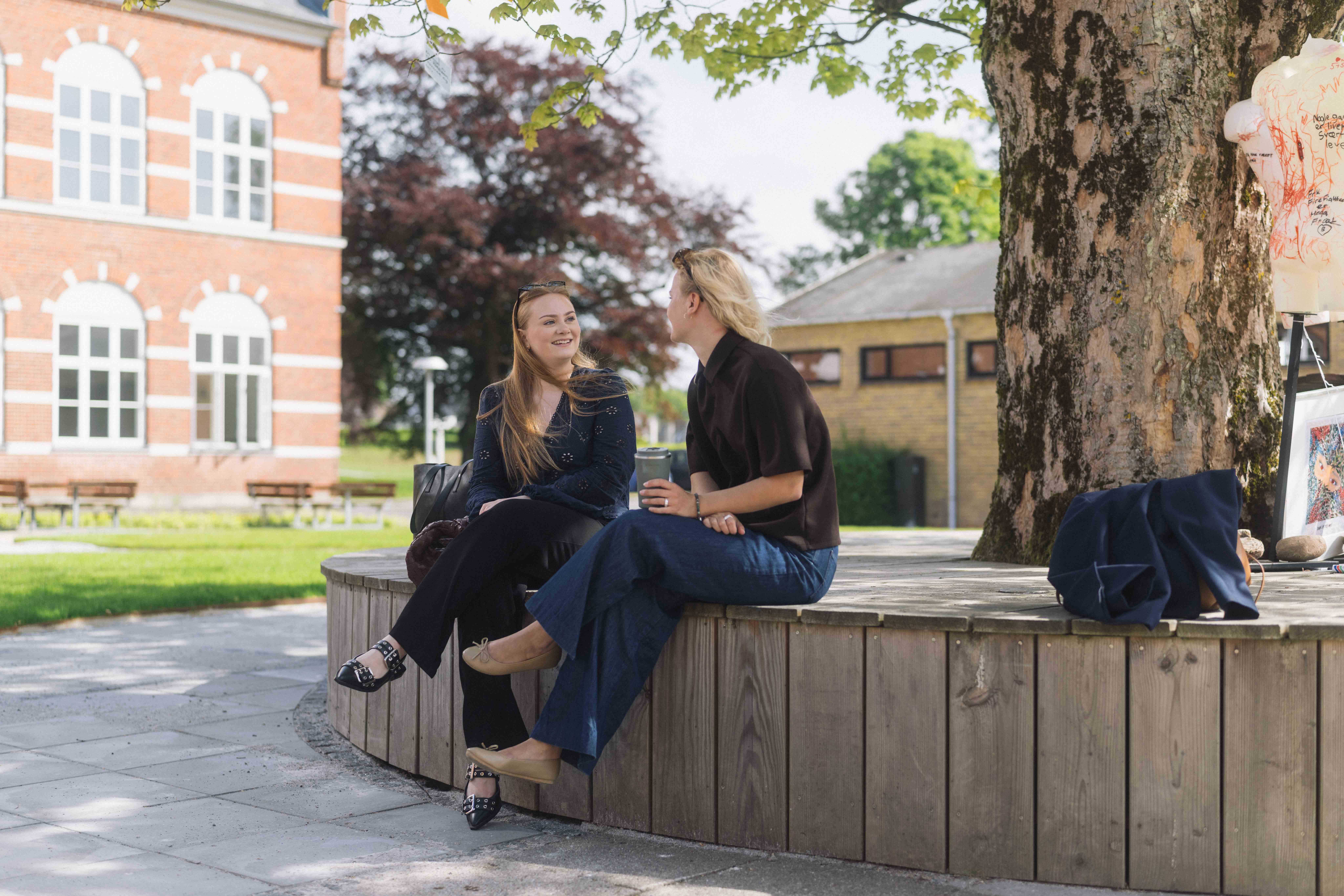 To unge kvinder sidder og snakker foran bygningerne til UCL Jelling.